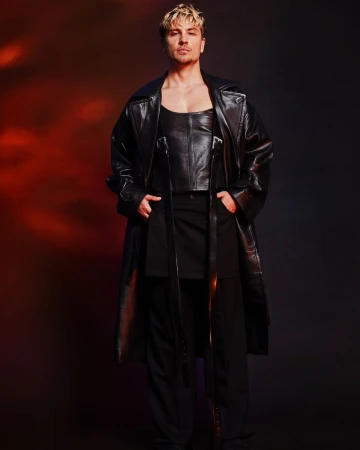 Adult male fashion model in black leather top and long coat poses in a studio with a red and black gradient background.