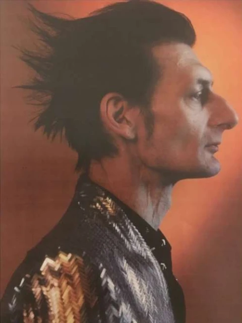 Profile portrait of an adult male with dark hair styled upward, wearing a patterned jacket against an orange backdrop.