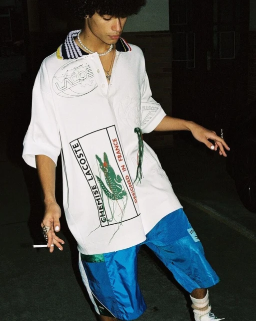 Person wearing Lacoste shirt and blue shorts stands outside. The image features a fashion style, with focus on the person's outfit.