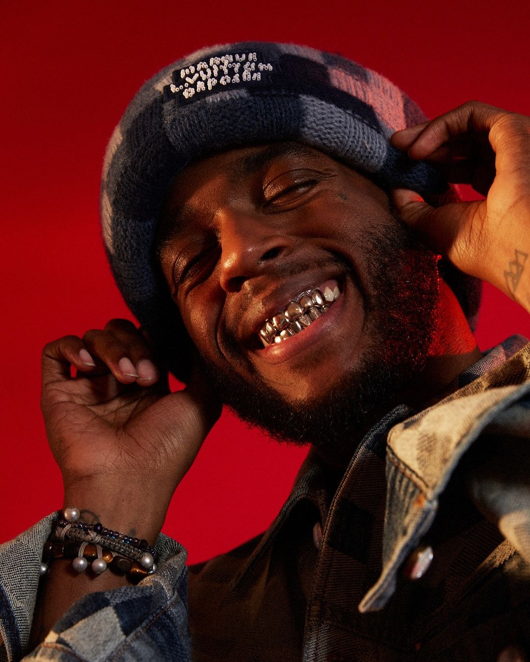 Close-up of smiling Black adult male wearing a patterned beanie, flannel jacket, and dental grillz against a red background.