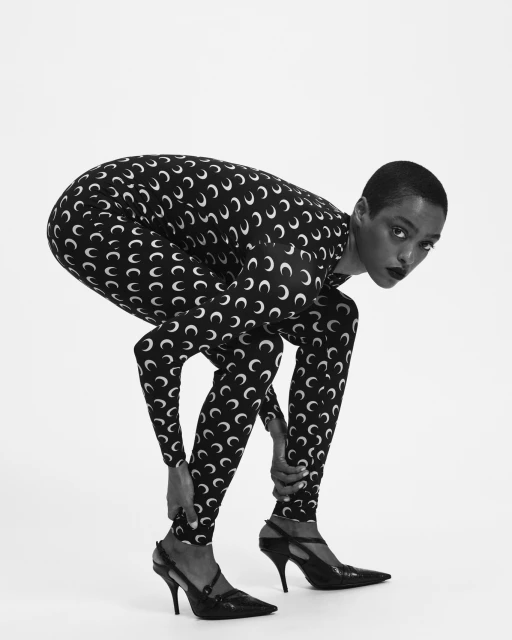 Black and white studio shot of a woman in patterned catsuit and heeled shoes bending over with hands on ankles, looking at the camera.