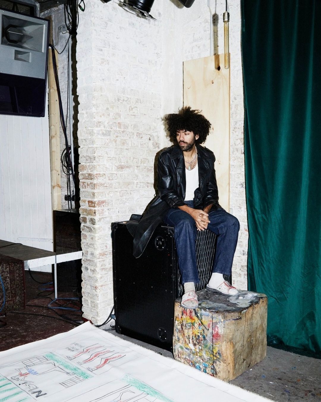 Bearded man with afro sits in indoor setting wearing a leather jacket, white undershirt, and jeans, with a green curtain backdrop.