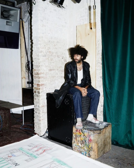 Bearded man with afro sits in indoor setting wearing a leather jacket, white undershirt, and jeans, with a green curtain backdrop.