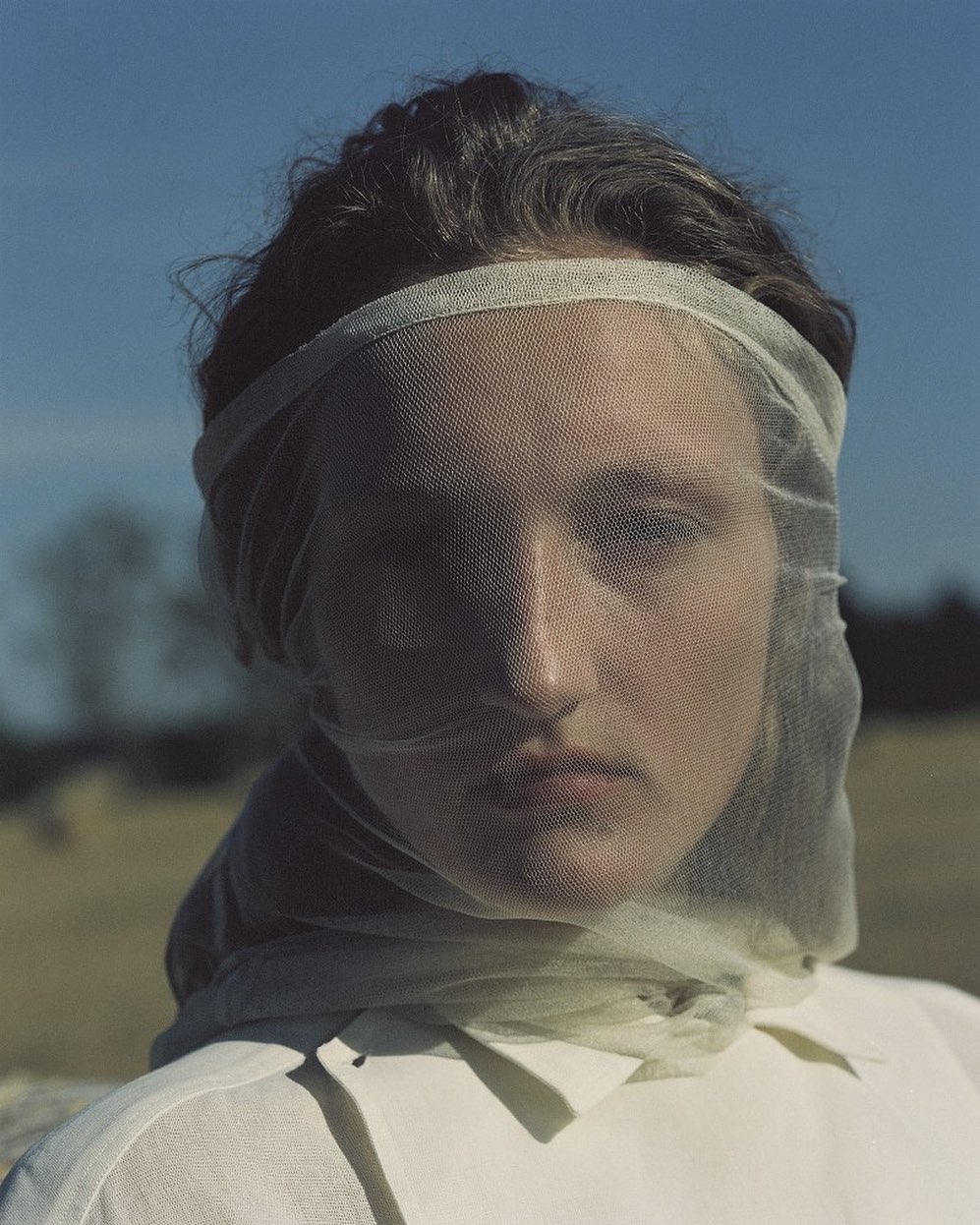 Close-up portrait of a woman with dark hair wearing a sheer veil and a white collared shirt, standing outdoors on a sunny day.
