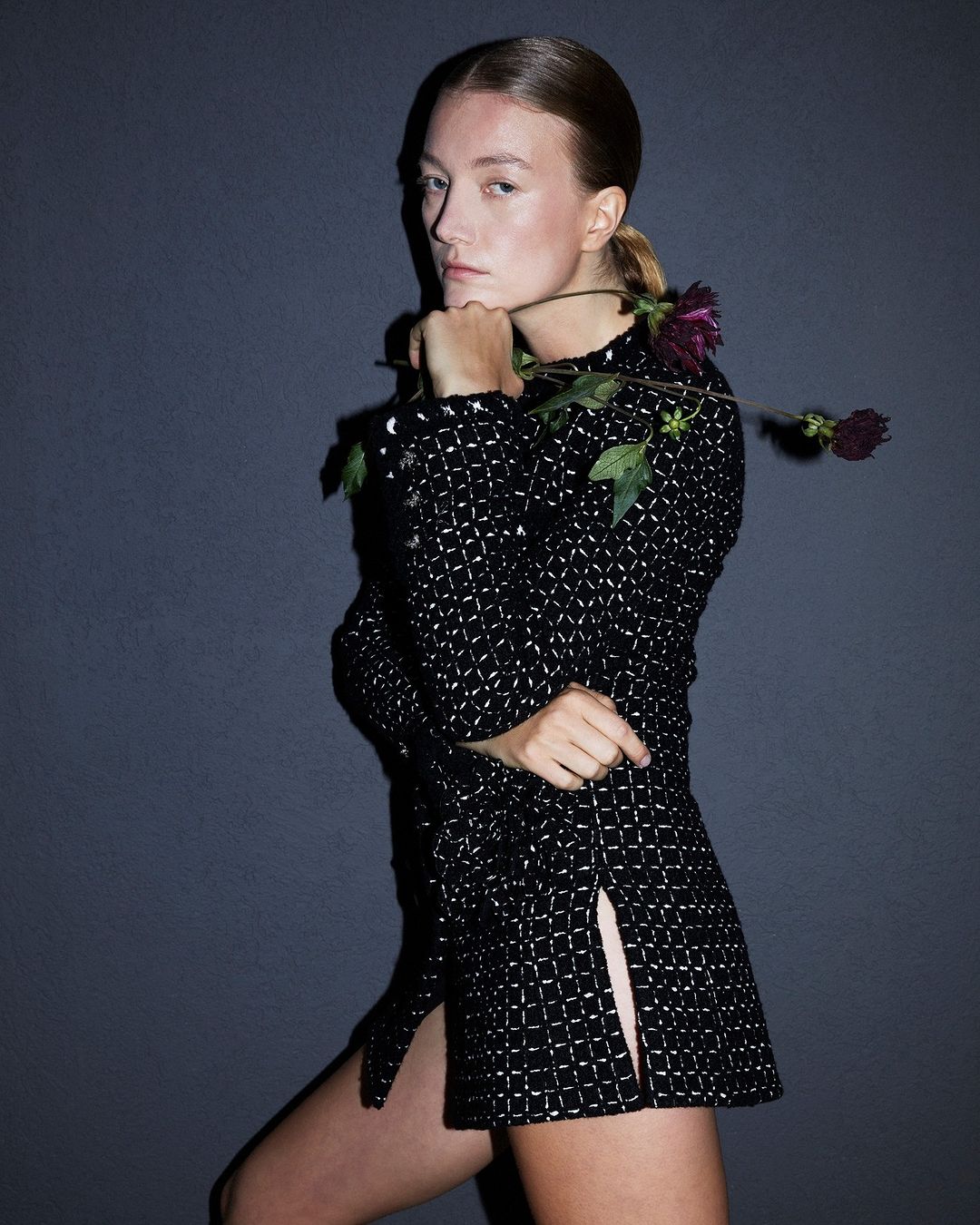 Portrait of a young woman with fair skin and auburn hair in a black and silver patterned dress, holding a dark red flower against gray background.