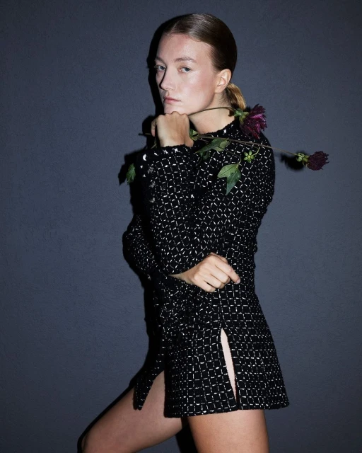 Portrait of a young woman with fair skin and auburn hair in a black and silver patterned dress, holding a dark red flower against gray background.