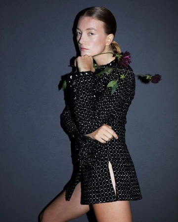 Portrait of a young woman with fair skin and auburn hair in a black and silver patterned dress, holding a dark red flower against gray background.