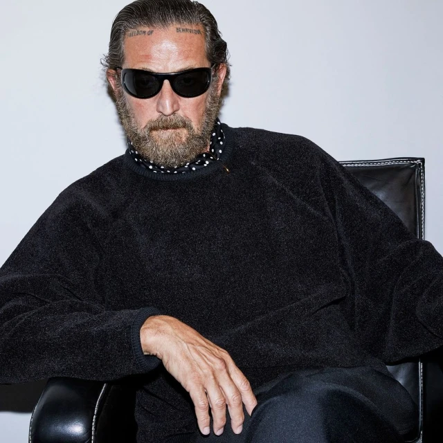 Portrait of a bearded adult male with sunglasses wearing a black sweater, captured indoors against a simple grey backdrop, seated in a chair.