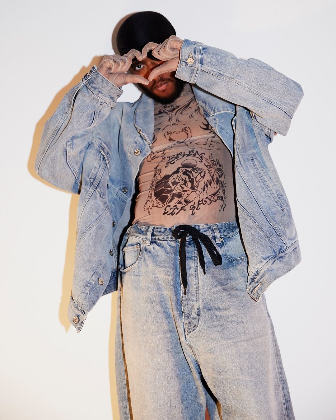Man wearing a denim jacket and mesh top posing with hands in a heart shape against a plain background