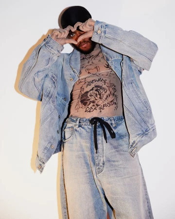 Man wearing a denim jacket and mesh top posing with hands in a heart shape against a plain background