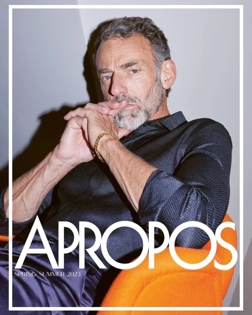 Close-up of a mature man with grey hair and a short beard, wearing a dark patterned shirt and gold bracelet, sitting in an orange chair.