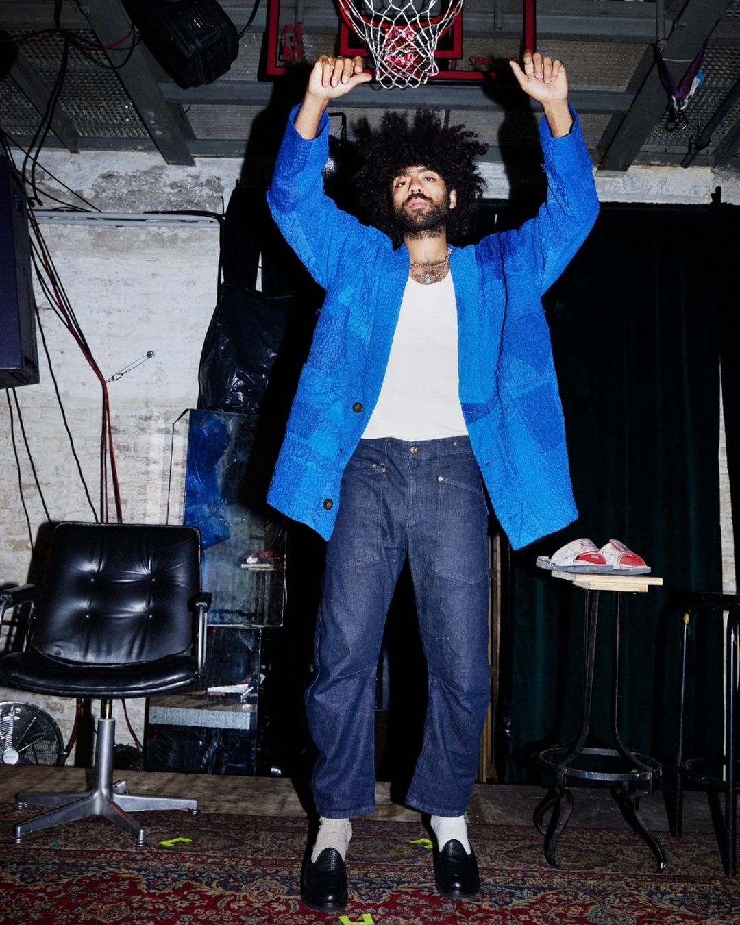 A man with an afro in a blue jacket and dark jeans stands in a casual indoor setting, raising his arms towards a basketball hoop.