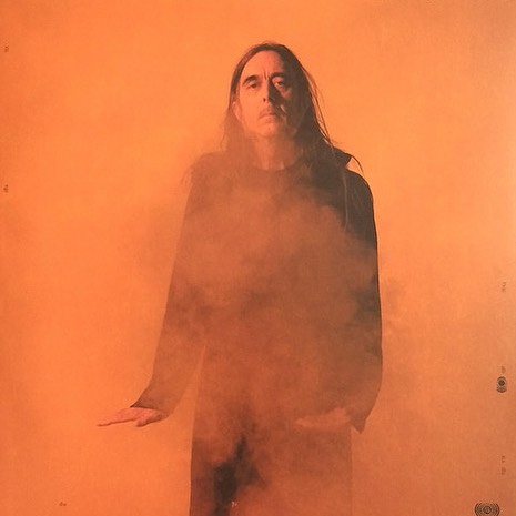 Adult male standing in front of an orange backdrop with smoke effect. The man has long hair and is wearing a dark shirt.