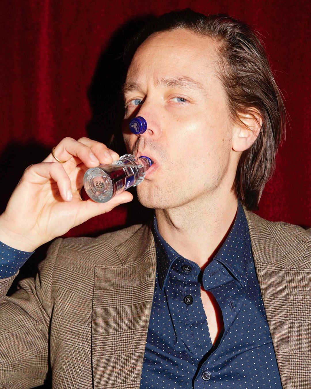 Adult male in a patterned shirt and blazer drinks from a short glass bottle with a cap on his nose against a blurred red background.