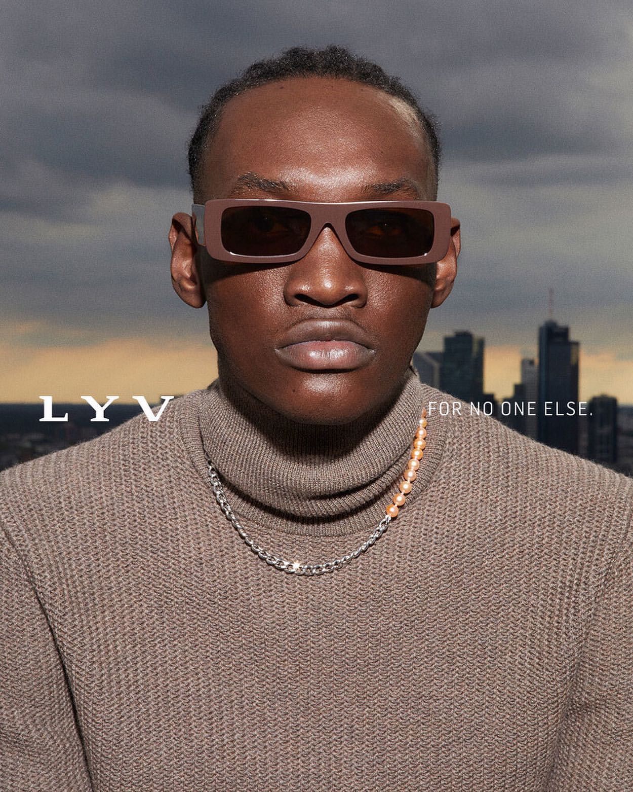 Portrait of a dark-skinned male wearing brown sunglasses and a turtleneck sweater, posing in front of a city skyline and cloudy sky.