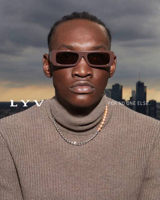 Portrait of a dark-skinned male wearing brown sunglasses and a turtleneck sweater, posing in front of a city skyline and cloudy sky.