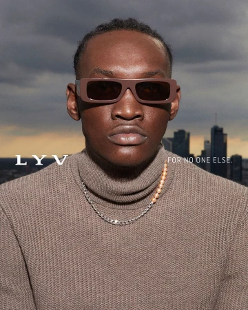 Portrait of a dark-skinned male wearing brown sunglasses and a turtleneck sweater, posing in front of a city skyline and cloudy sky.