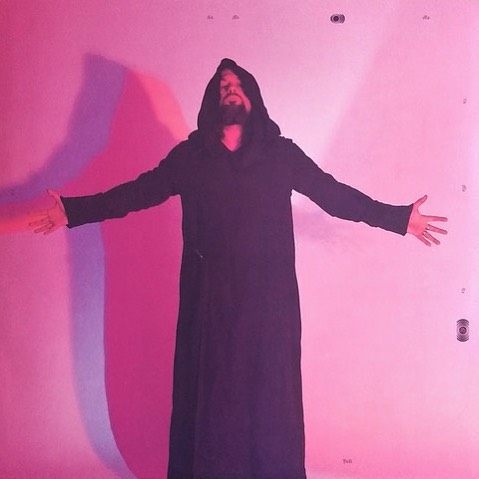 Man in black hooded robe stands with arms outstretched against a gradient pink background in a medium indoor studio shot.