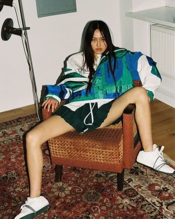 Young Asian woman with long dark hair sits in a chair wearing a color-block jacket, green shorts, and white sneakers in an indoor setting.