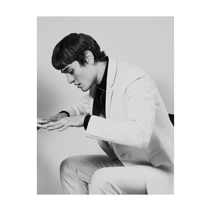 Monochromatic portrait of a young man in a white suit sitting with hands outstretched in an indoor setting, captured in a studio shot.