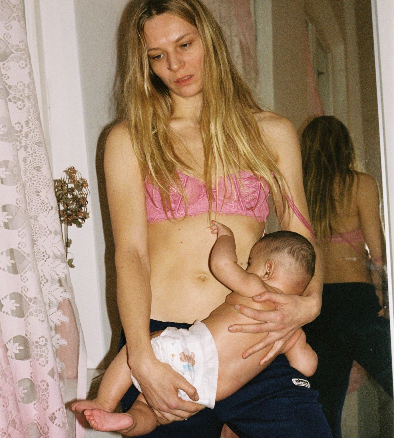 Indoor portrait of an adult female in a pink bra holding a baby in a diaper. The woman is standing, and the baby is lying in her arms.