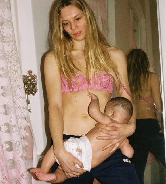 Indoor portrait of an adult female in a pink bra holding a baby in a diaper. The woman is standing, and the baby is lying in her arms.