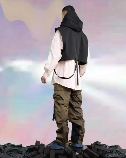Person from the back in a fashion editorial, wearing a black hooded vest, white shirt and cargo pants, stands on dark textured ground.