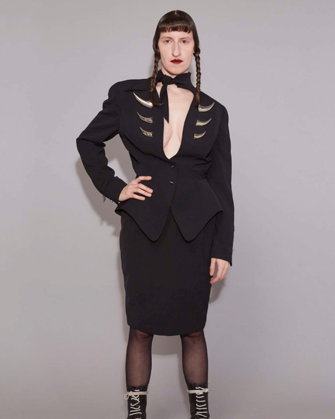 Woman in a black skirt suit with a deep V-neck and decorative silver elements, styled with braided hair, stands against a grey backdrop.