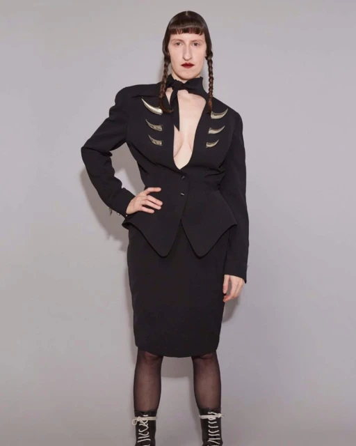 Woman in a black skirt suit with a deep V-neck and decorative silver elements, styled with braided hair, stands against a grey backdrop.