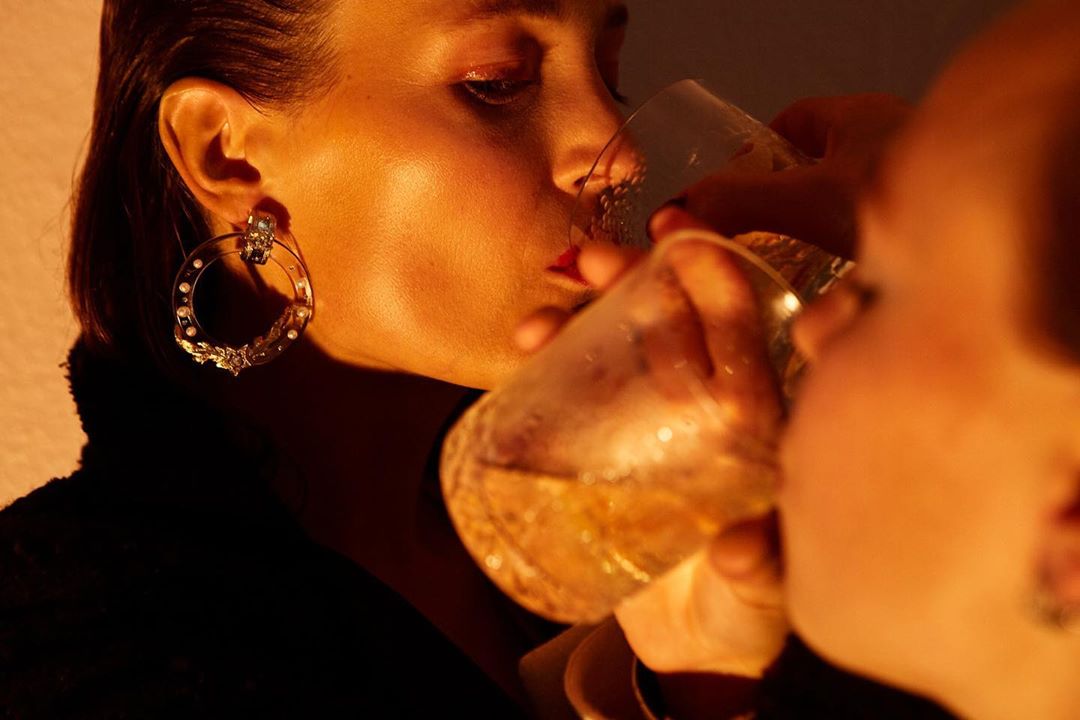 Close-up of a woman in warm amber light drinking from a glass, wearing a large statement hoop earring in Paris by Ronald Dick.