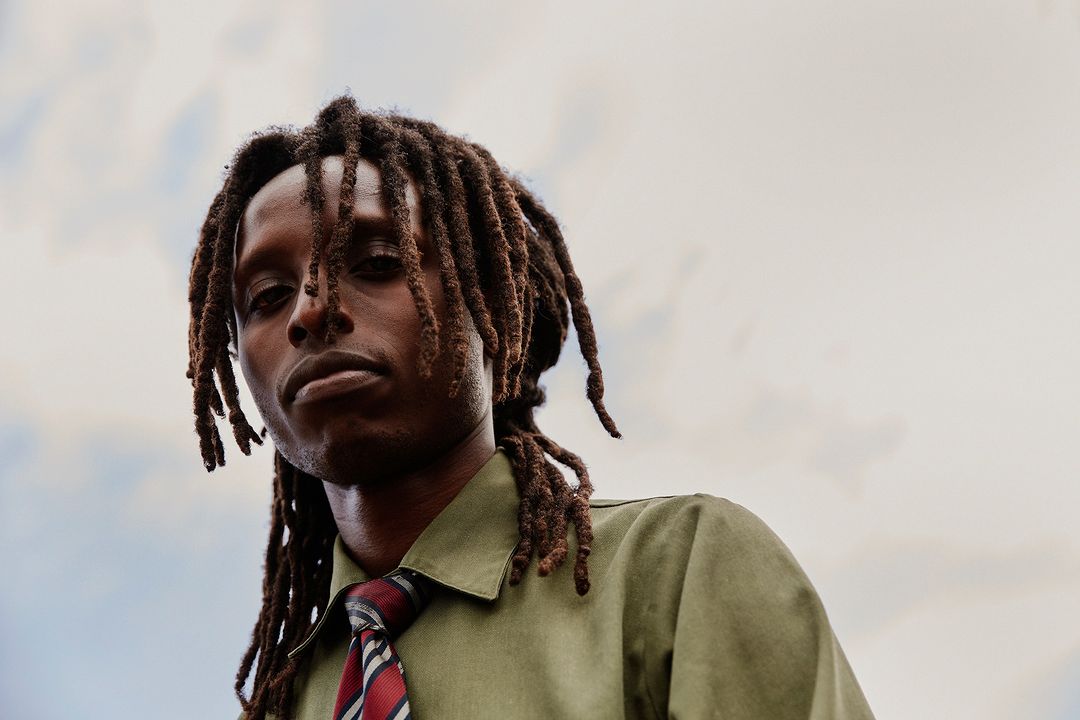 Portrait of an adult man with dreadlocks, wearing a green shirt and red striped tie, posing outdoors against a cloudy sky.