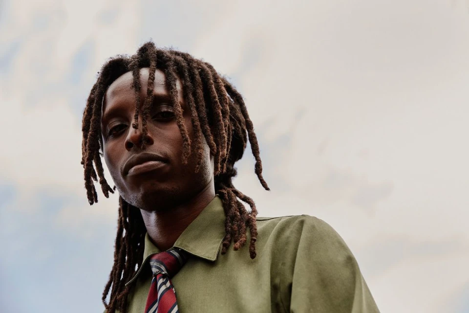 Portrait of an adult man with dreadlocks, wearing a green shirt and red striped tie, posing outdoors against a cloudy sky.