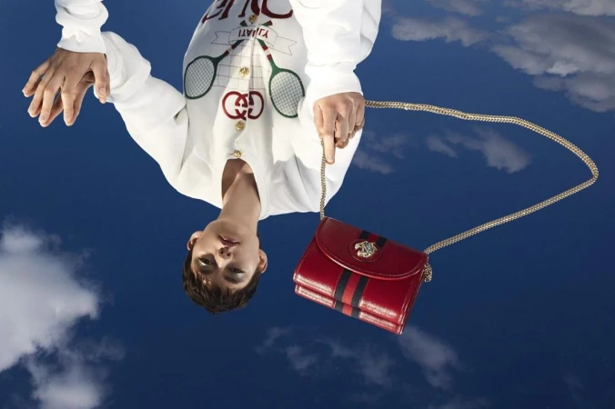 Upside-down view of a woman in white holding a red Gucci purse with a gold chain against a blue sky with clouds.