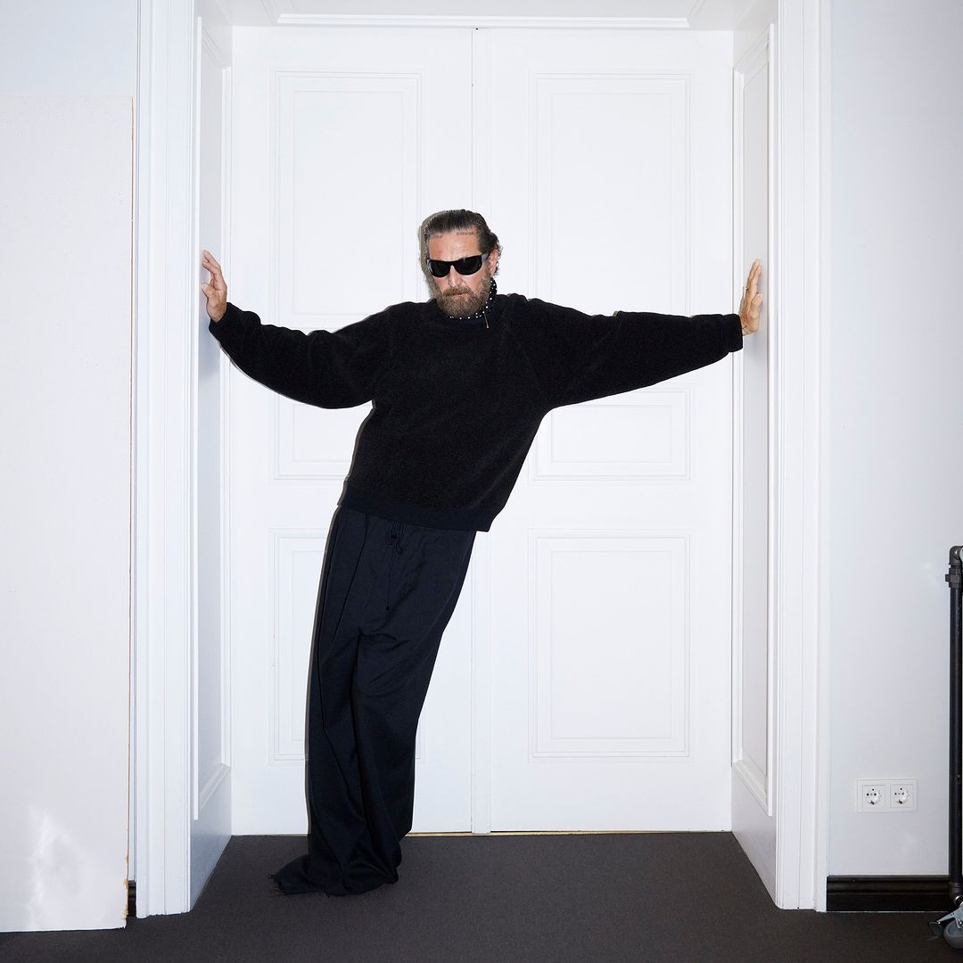Adult male with a beard, wearing sunglasses and dark clothing, leans against a white double door in an indoor setting.