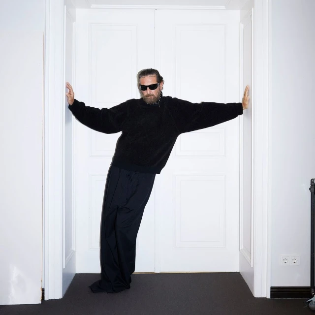 Adult male with a beard, wearing sunglasses and dark clothing, leans against a white double door in an indoor setting.