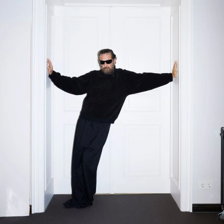 Adult male with a beard, wearing sunglasses and dark clothing, leans against a white double door in an indoor setting.
