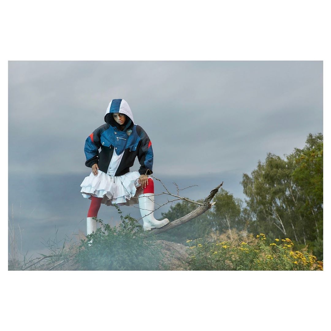 A woman in a hooded jacket, white skirt, and boots poses outdoors against an overcast sky, creating an edgy fashion-forward image.