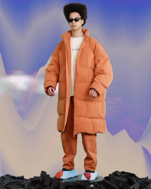A young adult male with an afro wearing a puffy orange coat and pants poses on a dark surface against a blurred, abstract cloud background.