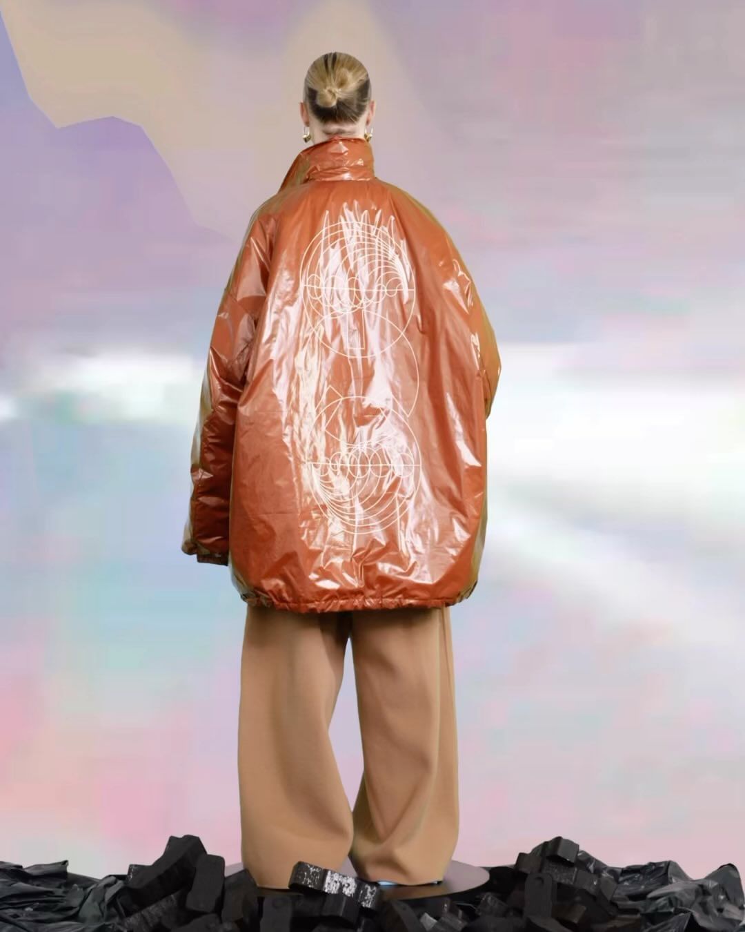 Back view of a slender adult female with hair in a bun, modeling an oversized orange puffer jacket with abstract designs, and wide-leg pants.