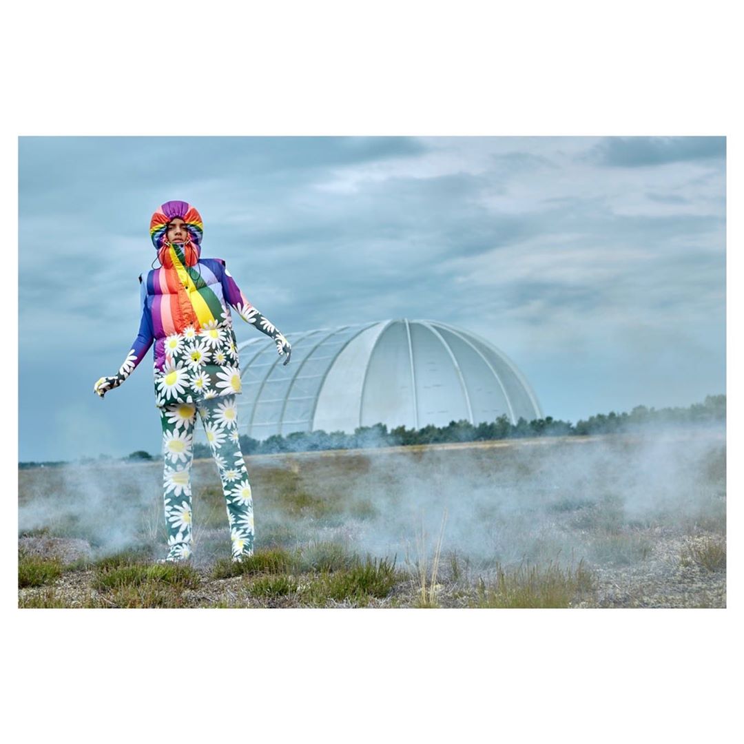 Person in rainbow and daisy print outfit stands outside with a geodesic dome structure in the background on an overcast day.