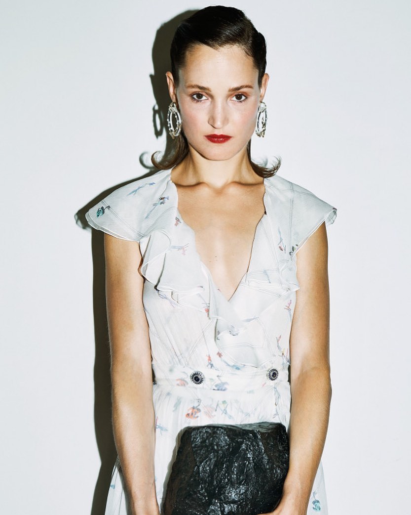 A fair-skinned woman with slicked-back brown hair and large earrings poses in a white dress, holding a black clutch against a stark white backdrop.