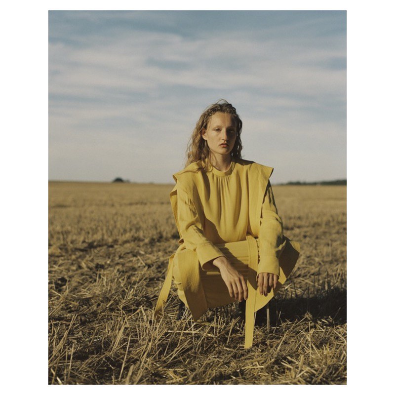 Woman in yellow outfit squats in a field. Fashion shot with natural light and rural setting. Fashion editorial photo featuring a model in nature.