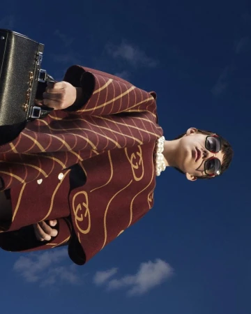 Model in patterned burgundy coat wearing oversized sunglasses holding a black handbag against clear blue sky