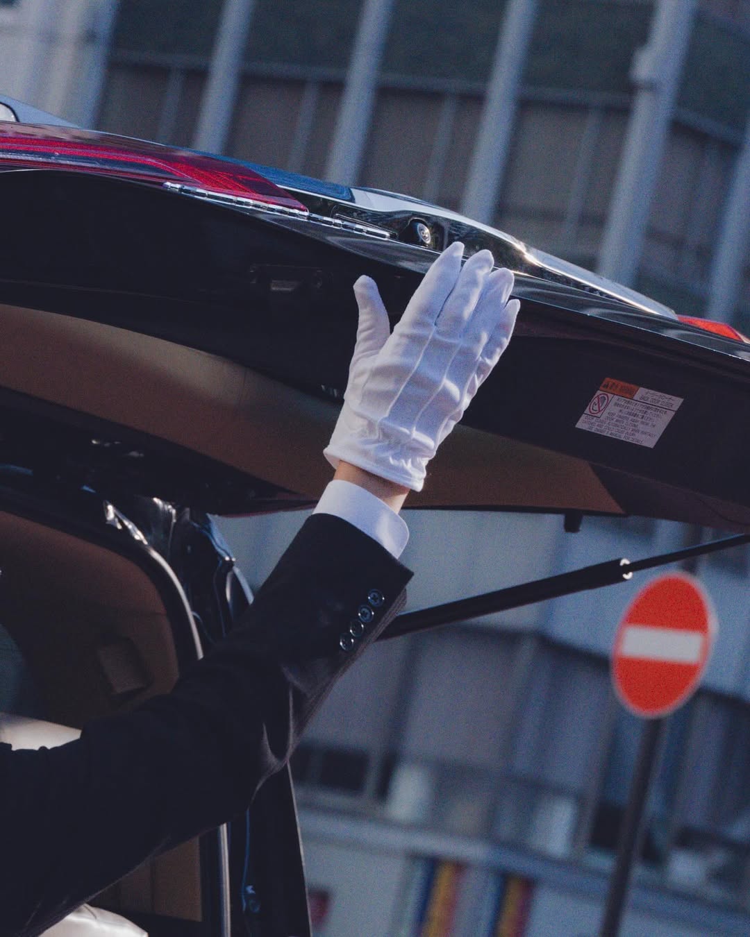 A gloved hand reaches toward an open car trunk in an urban setting, capturing a sense of service and luxury automotive transport.