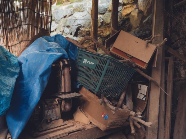 Pile of discarded items in storage area with a blue tarp, wooden planks, cardboard boxes, green plastic basket, and weathered equipment.
