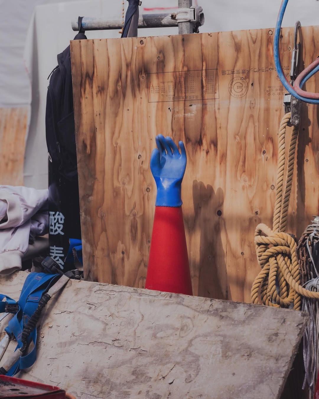 A blue and red protective sleeve stands in front of a plywood board with a rope, harnessing, and other equipment in the background.