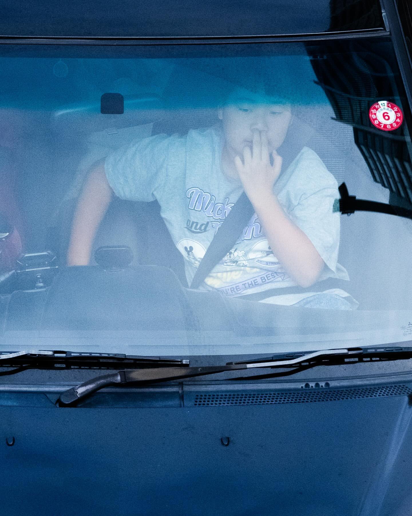Asian child in a car wearing a seatbelt and t-shirt with hand near mouth, car windshield and dashboard visible.