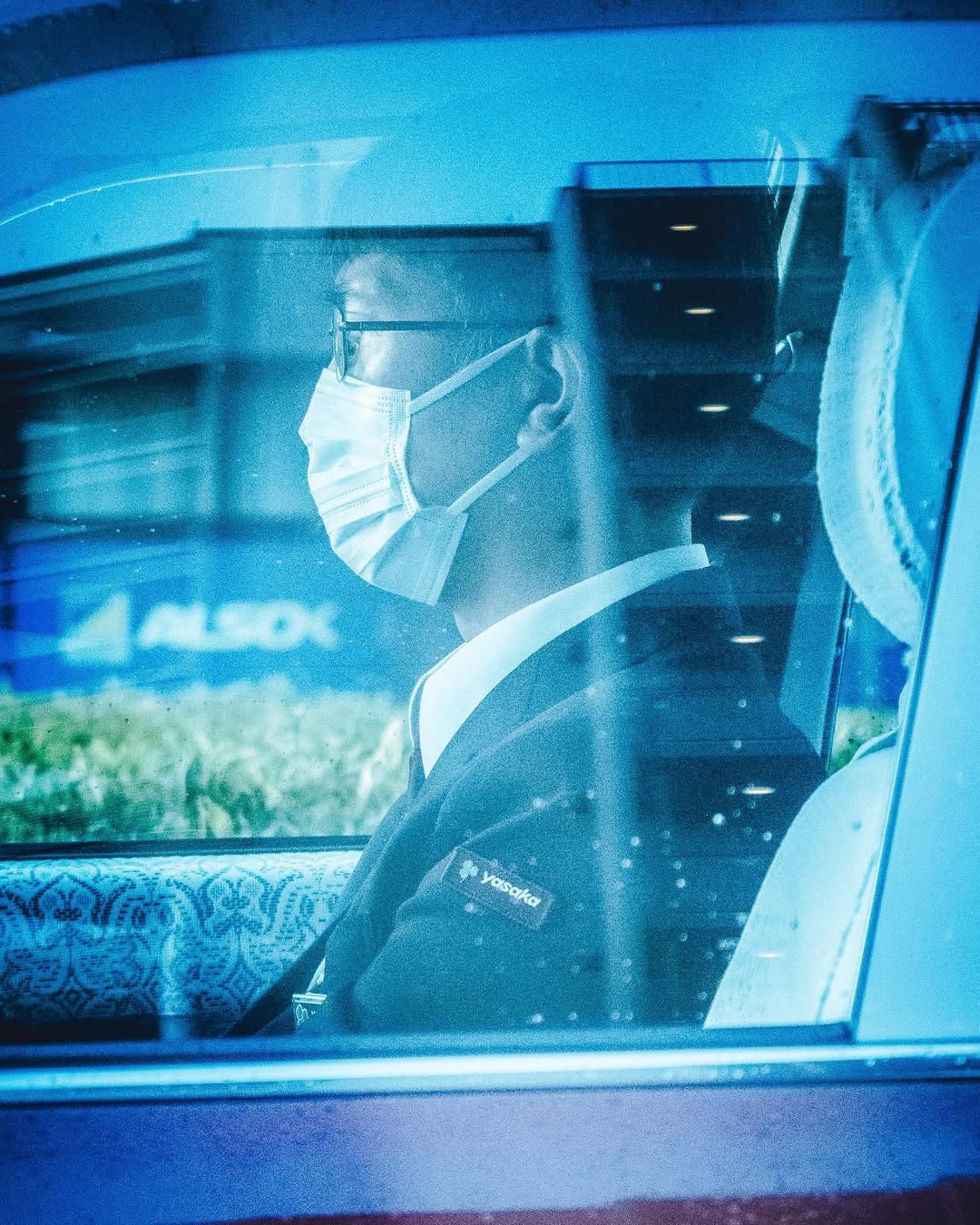 Adult male in a suit wearing glasses and a face mask is visible through the side window of a car in an urban setting.