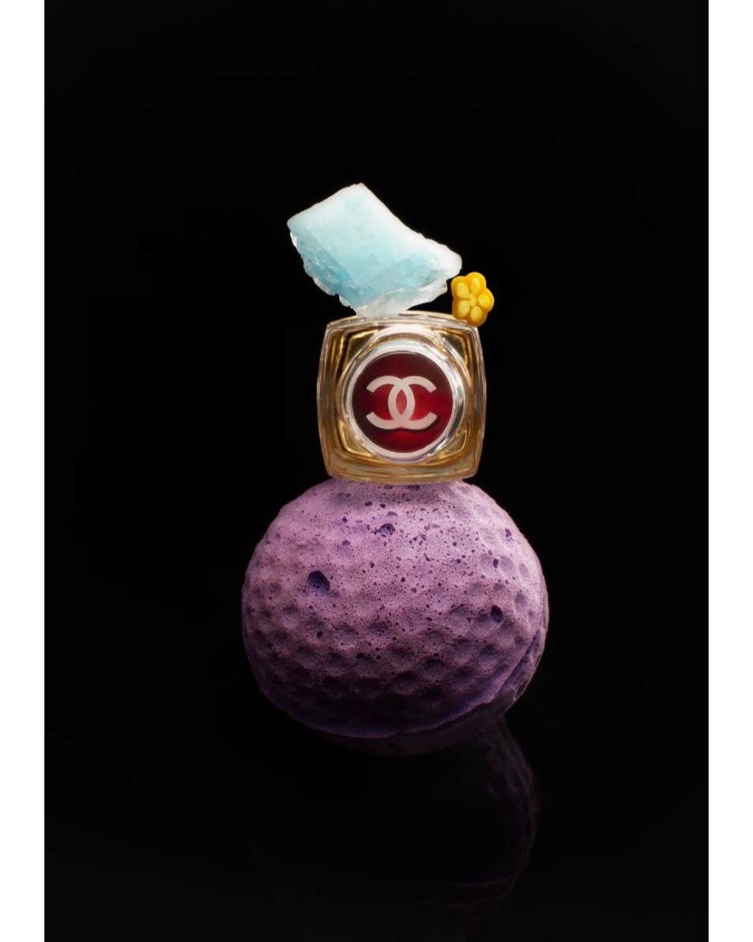 Chanel eye shadow pot on a purple sponge ball, balanced with a blue rock and yellow flower against a black background in a studio setting.