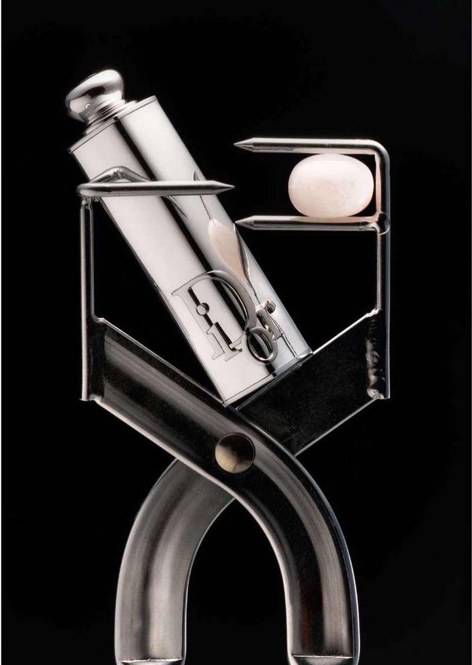 High-key product shot of a silver Dior tube held in a silver tool and a small, round pink stone against a black background.